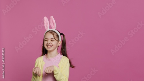 фотографія Joyful carefree schoolgirl jumping around in studio, imitating a rabbit and hopping against pink background