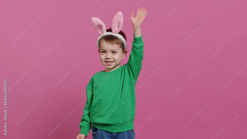 Joyful small kid waving and greeting someone in studio, smiling against pink background. Lovely little boy wearing fluffy bunny ears on his head, celebrating easter holiday. Camera A. Joyful small kid waving and greeting someone in studio, smiling against pink background. Lovely little boy wearing fluffy bunny ears on his head, celebrating easter holiday. Camera A.