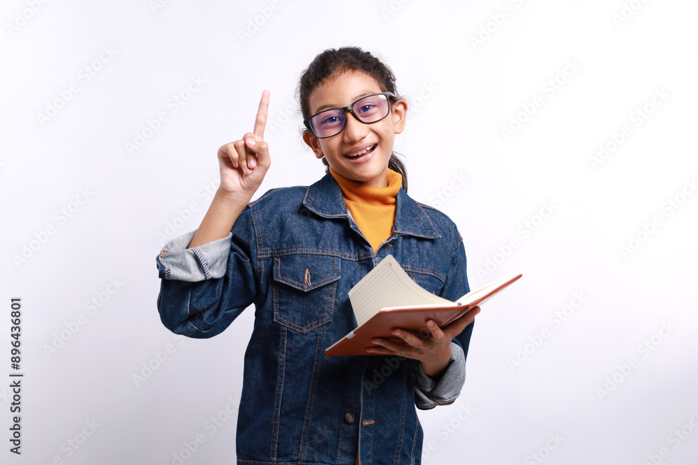 Smart kid in casual clothes and eyeglasses holding book and pointing index finger on white background. Aha moments, getting idea Smart kid in casual clothes and eyeglasses holding book and pointing index finger on white background. Aha moments, getting idea