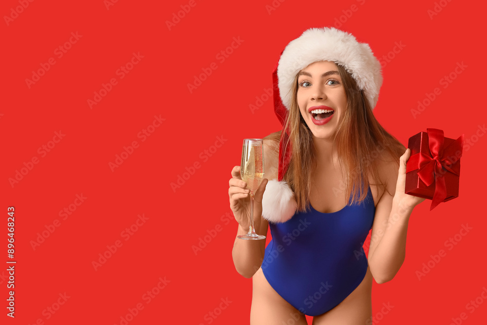 Happy young woman in swimsuit and Santa hat holding glass of champagne with gift box on red background. Christmas in July Happy young woman in swimsuit and Santa hat holding glass of champagne with gift box on red background. Christmas in July