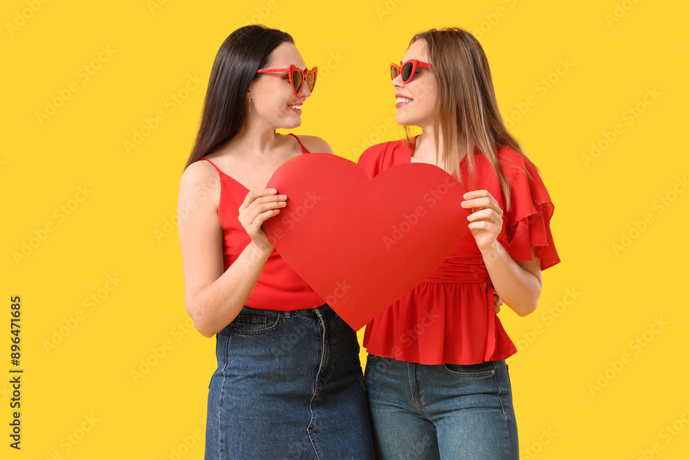 Beautiful young lesbian couple with paper heart on yellow background Beautiful young lesbian couple with paper heart on yellow background