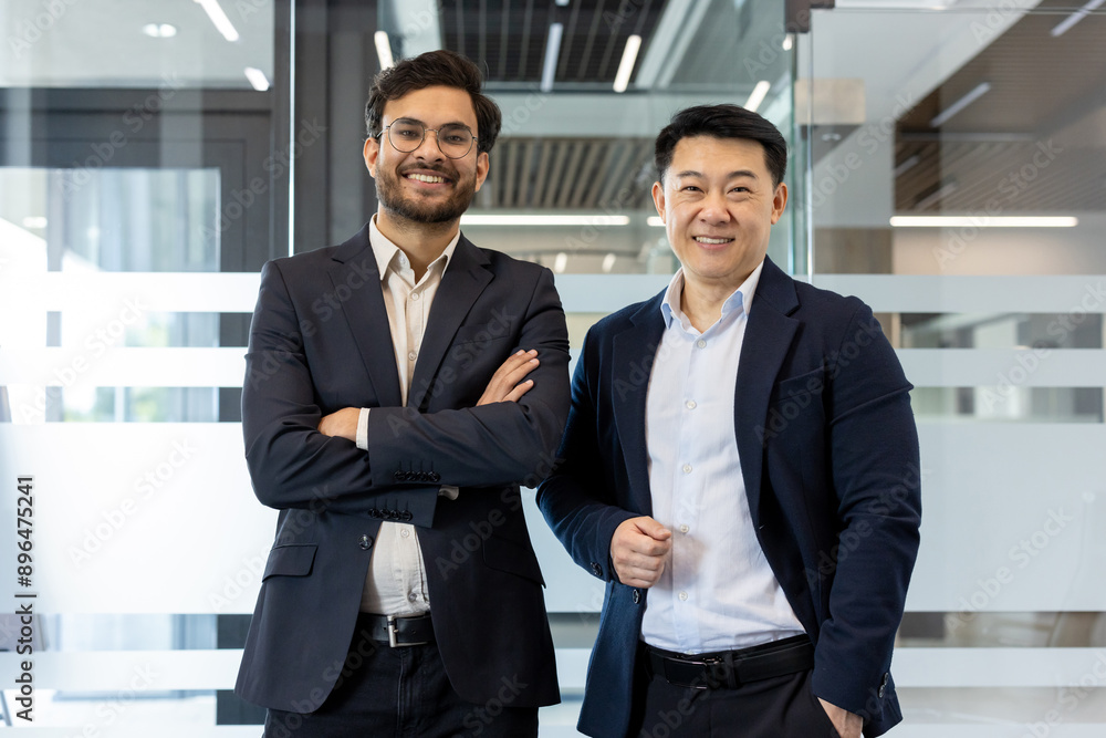 Two confident businessmen standing in modern office. One with arms crossed and other with hands in pockets. They express teamwork, professionalism, and success in business environment. Two confident businessmen standing in modern office. One with arms crossed and other with hands in pockets. They express teamwork, professionalism, and success in business environment.