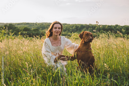 young cheerful woman playing with her dog outdoors in beautiful countryside field. Female and ridgeback dog walking and having fun