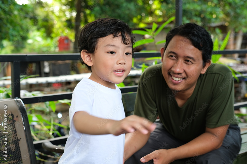 A young boy in a white shirt enthusiastically gestures while his father smiles supportively in the background. This image captures a candid moment of father-son bonding and communication, A young boy in a white shirt enthusiastically gestures while his father smiles supportively in the background. This image captures a candid moment of father-son bonding and communication,