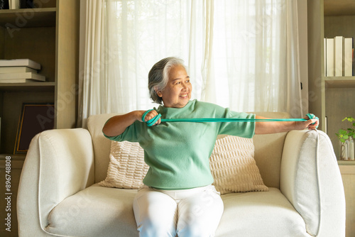 Happy Asian senior mature woman enjoy healthy lifestyle resistance band training exercise for stretching body muscle shoulder and arm on sofa in living room. Elderly self-care and home fitness concept photo