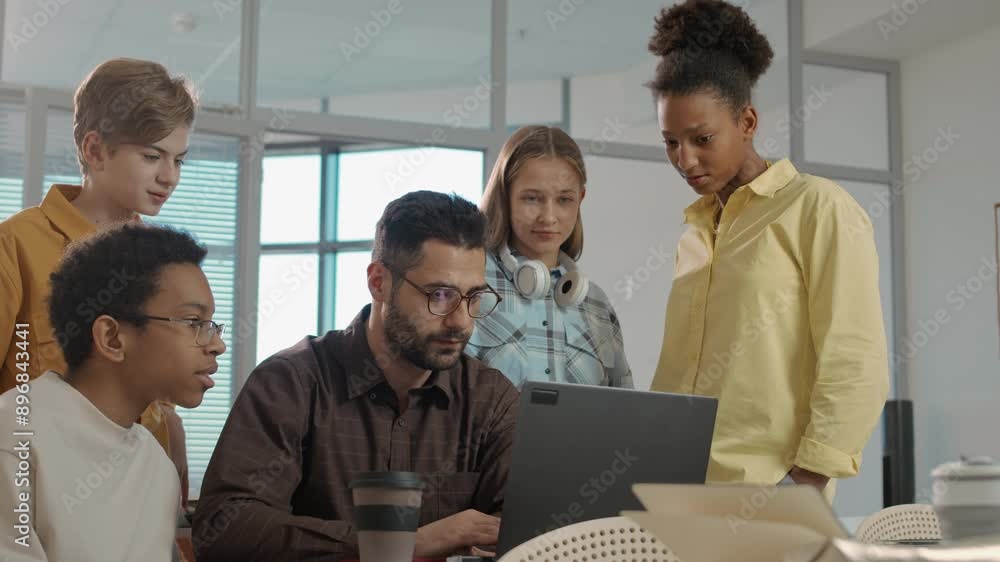 Medium shot of group of multiethnic teenage children gathering near young Caucasian male teacher sitting at desk with laptop, discussing collective educational project and sharing ideas Medium shot of group of multiethnic teenage children gathering near young Caucasian male teacher sitting at desk with laptop, discussing collective educational project and sharing ideas