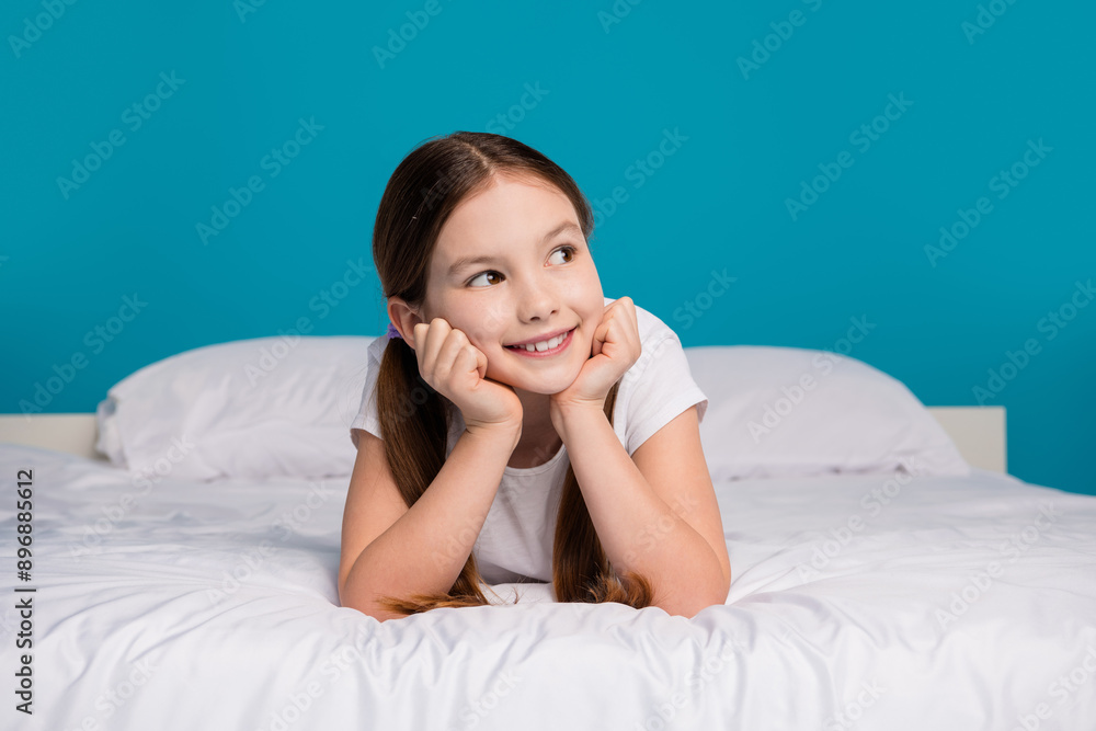 Full size photo of charming little girl look dreamy empty space lying bed awakening bedroom wear pajama isolated on blue color background Full size photo of charming little girl look dreamy empty space lying bed awakening bedroom wear pajama isolated on blue color background