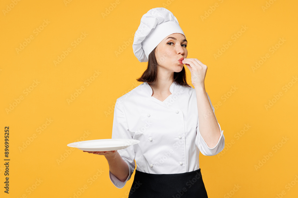 Young happy chef cook baker woman wear white shirt black apron uniform toque chefs hat hold empty plate making okay taste delight sign isolated on plain yellow background studio. Cooking food concept. Young happy chef cook baker woman wear white shirt black apron uniform toque chefs hat hold empty plate making okay taste delight sign isolated on plain yellow background studio. Cooking food concept.