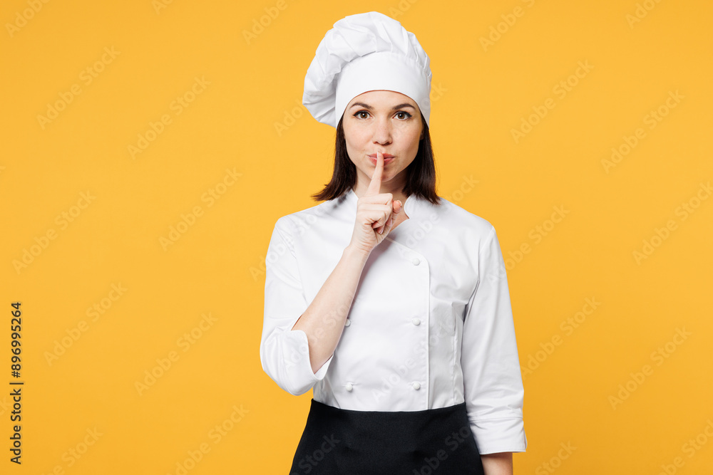Young chef cook baker woman wear white shirt black apron uniform toque chefs hat say hush be quiet with finger on lips shhh gesture isolated on plain yellow background studio. Cooking food concept. Young chef cook baker woman wear white shirt black apron uniform toque chefs hat say hush be quiet with finger on lips shhh gesture isolated on plain yellow background studio. Cooking food concept.