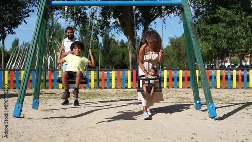 Latin family with afro hair enjoying a day at the playground. Father and mother pushing their son and baby on the swings
