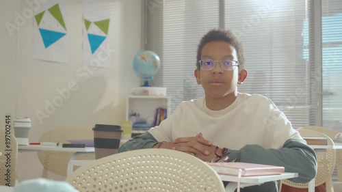 Portrait of serious teenage African American boy wearing glasses sitting at desk in classrom at school during recess time and looking at camera photo