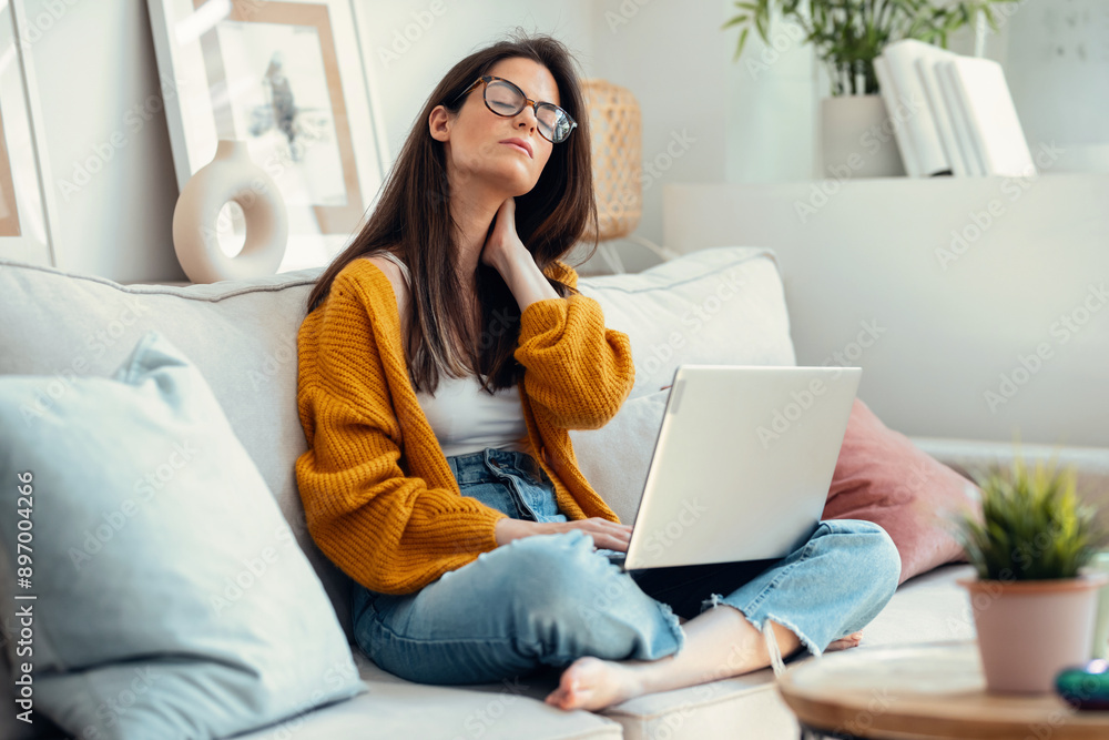 Tired young woman with neck pain using her laptop sitting on couch at home Tired young woman with neck pain using her laptop sitting on couch at home