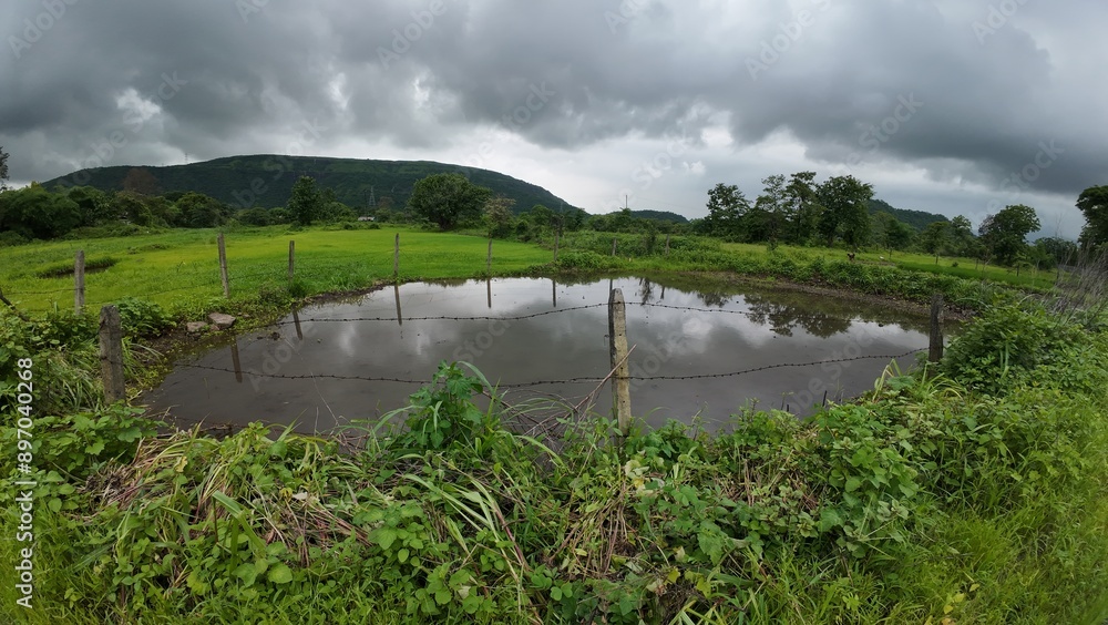 A serene water body bordered by a wired fence, lush green grass, and mountains, under a cloudy sky during monsoon.
A serene water body bordered by a wired fence, lush green grass, and mountains, under a cloudy sky during monsoon.