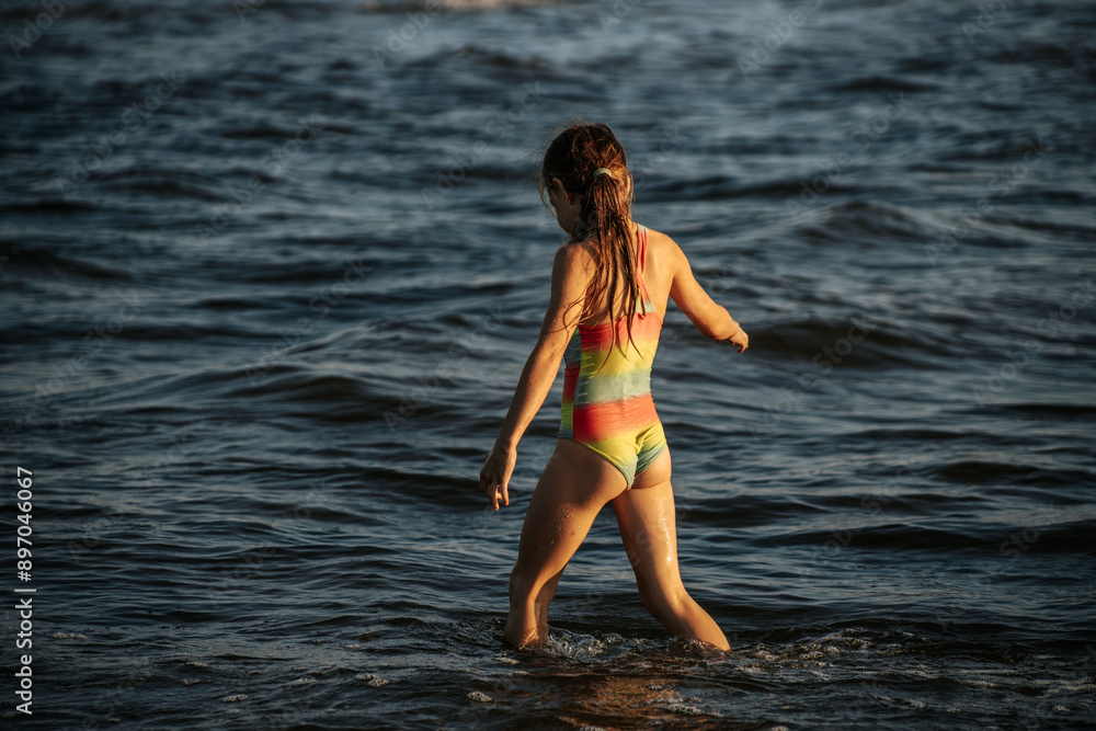 Fototapeta premium A girl in a colorful swimsuit playing energetically in the ocean waves, capturing a moment of joy and movement by the sea. The image emphasizes the fun and excitement of a summer day at the beach