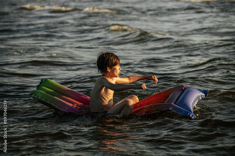 Obraz premium A boy riding an inflatable raft in the waves at the beach, capturing a moment of fun and adventure in the water. The image shows his determination and enjoyment as he navigates the waves.