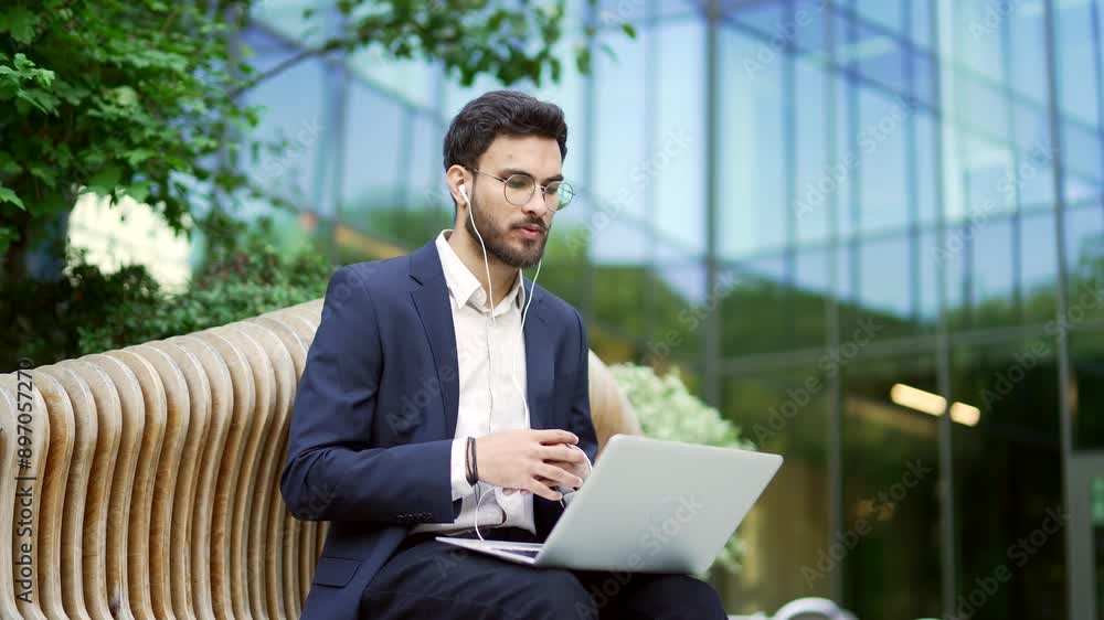 Businessman in suit talking on a video call using laptop while sitting on a bench near an office building. Confident entrepreneur in headphones having an online meeting with client or business partner Businessman in suit talking on a video call using laptop while sitting on a bench near an office building. Confident entrepreneur in headphones having an online meeting with client or business partner