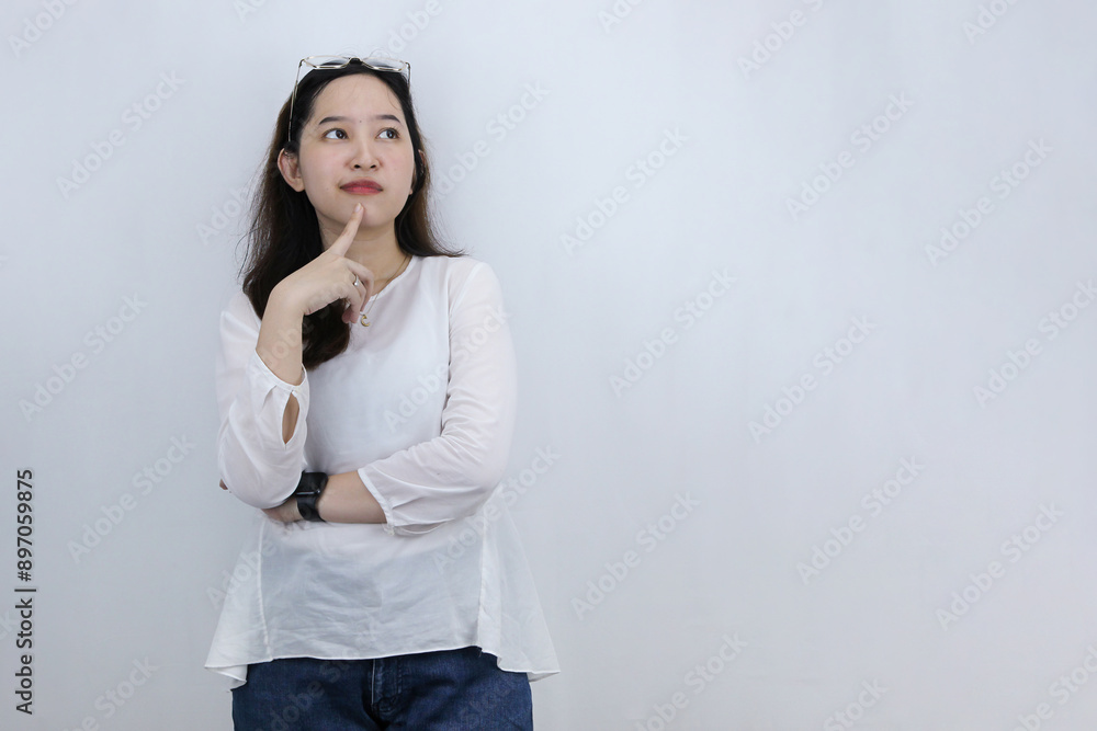 Asian woman keeps index finger on chin looks pensively above dressed in casual white shirt poses against white background, thinks deeply about something Asian woman keeps index finger on chin looks pensively above dressed in casual white shirt poses against white background, thinks deeply about something