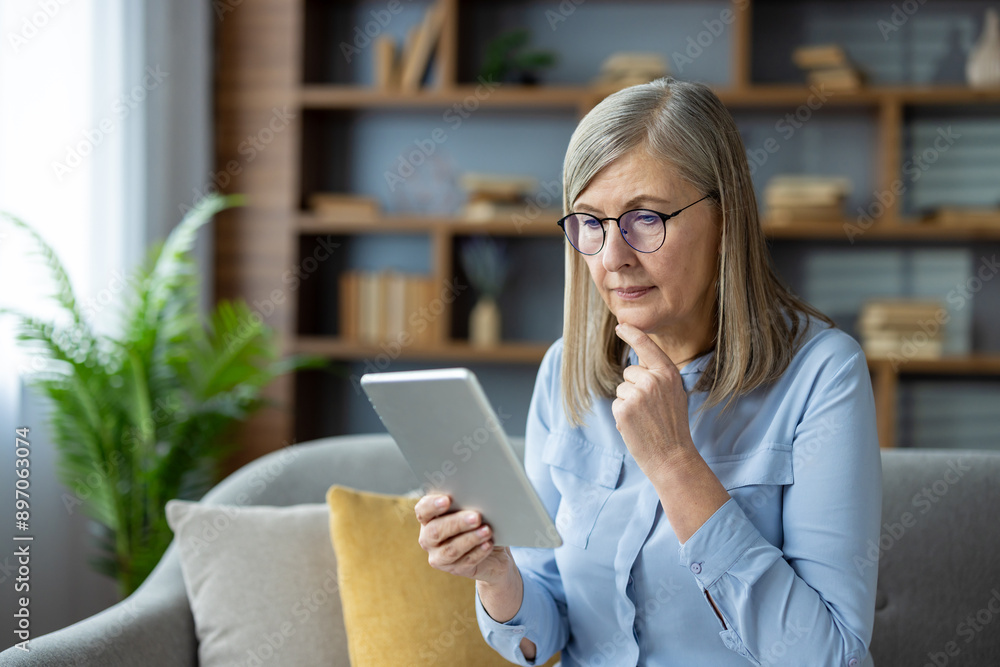 Senior woman with glasses reading on tablet in modern living room thoughtfully. She seems deeply engaged with information on the device showing concentration and curiosity. Senior woman with glasses reading on tablet in modern living room thoughtfully. She seems deeply engaged with information on the device showing concentration and curiosity.