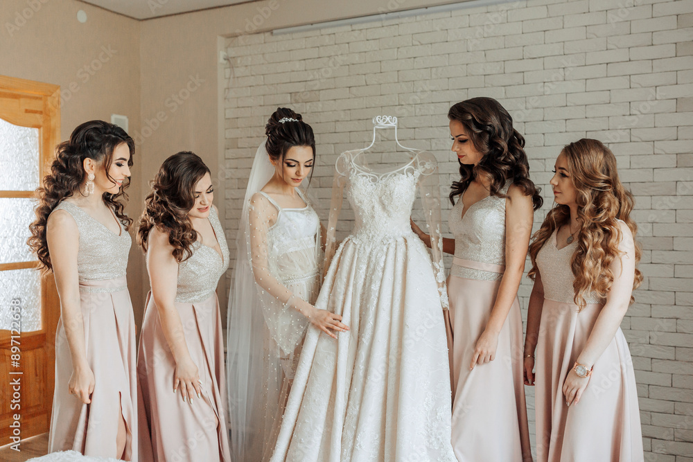 A group of women are standing around a wedding dress. The bride is looking at the dress and the other women are looking on. Scene is one of excitement and anticipation for the upcoming wedding