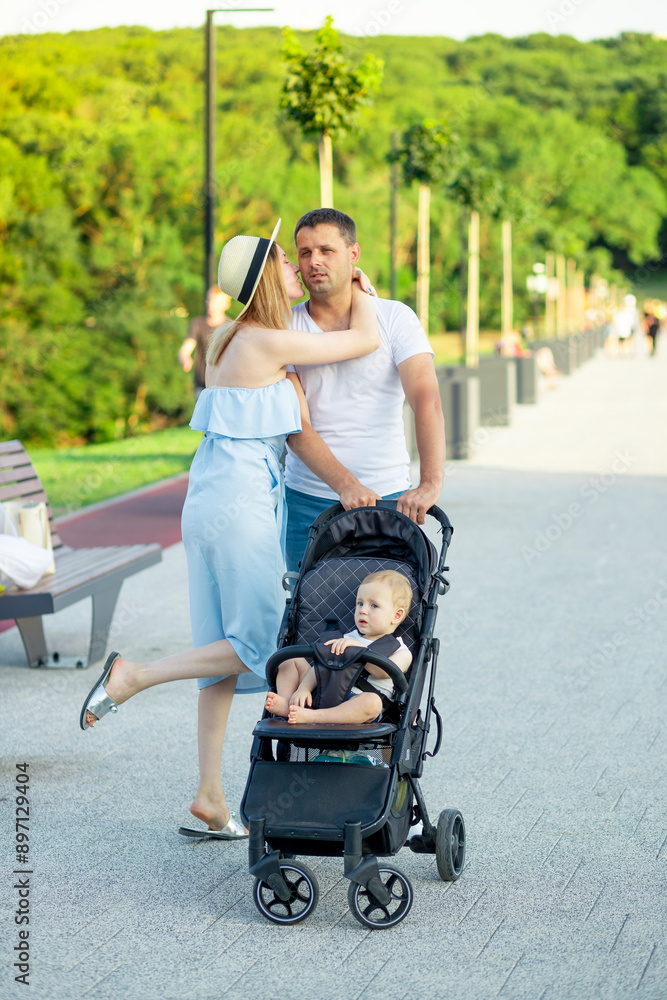 happy young parents mom and dad are walking with a baby in a stroller in the park in a hat in the summer in the setting sun and smiling having fun happy young parents mom and dad are walking with a baby in a stroller in the park in a hat in the summer in the setting sun and smiling having fun