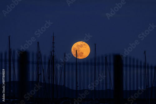 24 FEBRUARY 2024 wolf moon rising over the harbour at Weymouth, Dorset, UK, blue sky, pink moon, boat masts and fence photo
