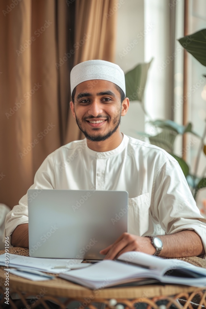 A young man wearing a hijab smiles at the camera while working on his laptop in a home office. His attire and the modern technology suggest an inclusive, contemporary work environment. A young man wearing a hijab smiles at the camera while working on his laptop in a home office. His attire and the modern technology suggest an inclusive, contemporary work environment.