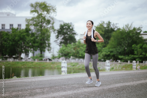 A young woman jogs in the city on a summer morning, embodying an active lifestyle. This fit athlete, dressed in sports clothing, enjoys outdoor exercise, promoting health, vitality, and a healthy life © Witoon