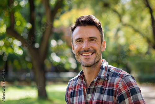 A young man with brown hair and beard, wearing a plaid shirt, smiling and standing on the street, yellow-green blurred background