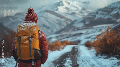 A backpacker bundled in warm clothing stands on a snowy path, looking towards the breathtaking, mountainous scenery covered in snow under a cloudy sky. photo