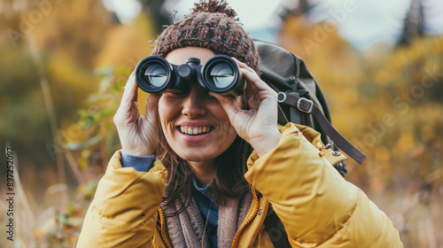 A smiling woman in a beanie birdwatching with binoculars in an autumnal setting, emphasizing enjoyment and connection with nature.