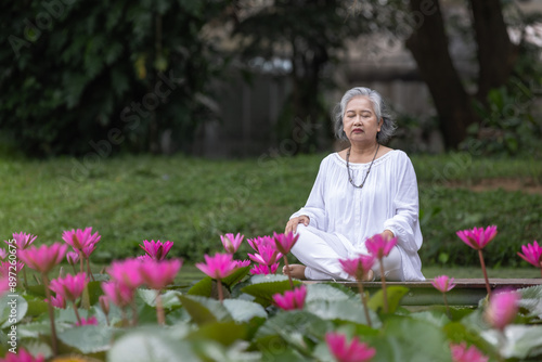 Elderly Woman Meditating by Pond Surrounded by Blooming Pink Lotus Flowers in a Tranquil Park photo