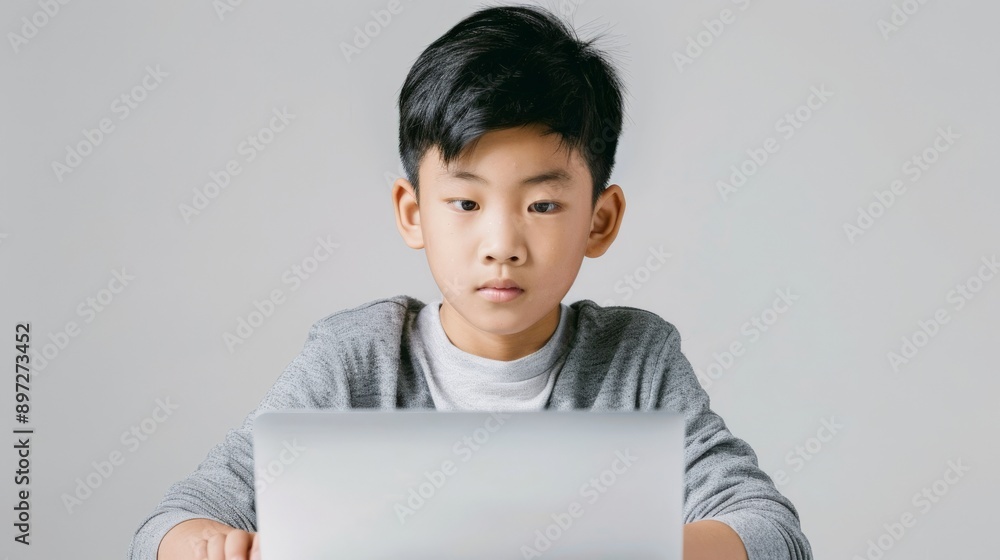 Asian boy wearing grey long-sleeved shirt, using laptop, with focused serious expression, image is taken in minimalist studio setting with pure white backdrop, moment learning with technology. Asian boy wearing grey long-sleeved shirt, using laptop, with focused serious expression, image is taken in minimalist studio setting with pure white backdrop, moment learning with technology.