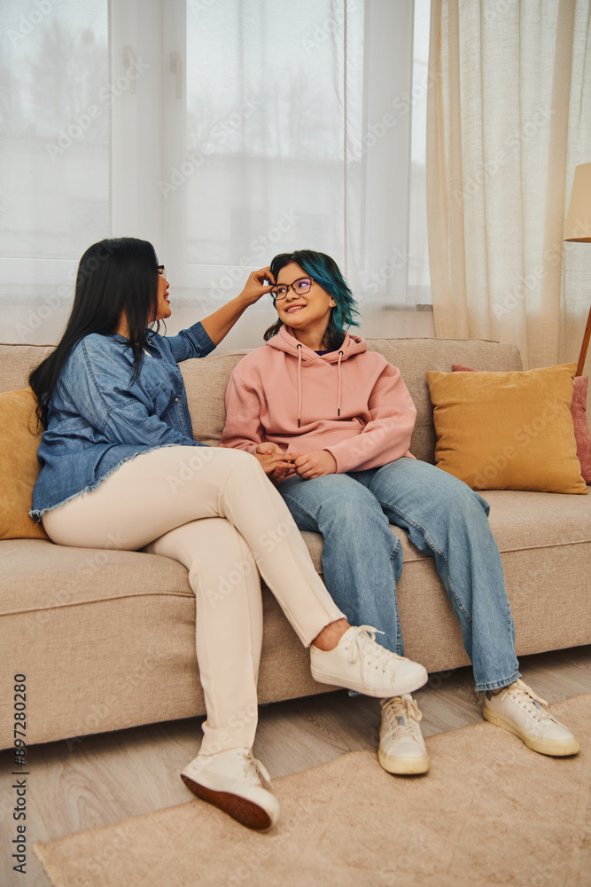 Asian mother and daughter in casual attire engaged in a lively conversation while sitting on a cozy couch at home.