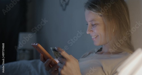 Happy young woman using smartphone and holding credit card paying in online store shopping sitting in bed in apartment in evening. E-commerce and money concept.