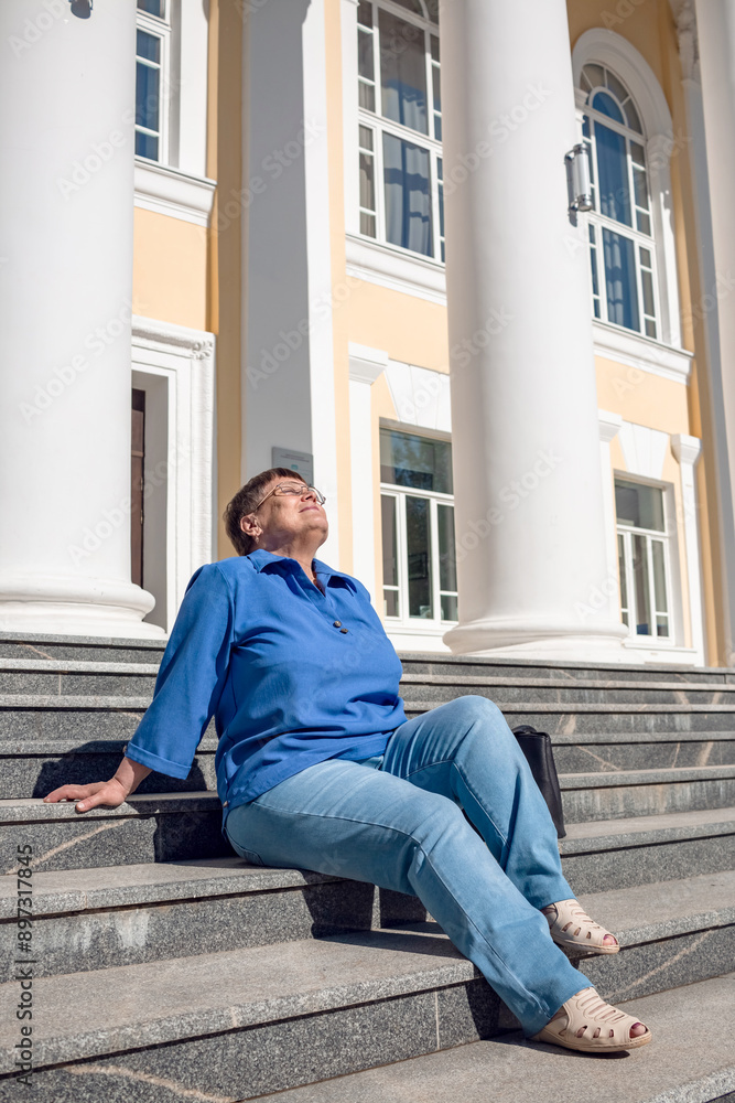 An elderly woman sits on the steps of a large white building with columns. An elderly woman sits on the steps of a large white building with columns.