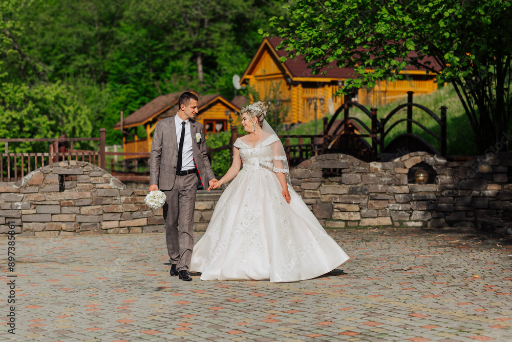 Fototapeta premium A bride and groom are walking down a path in front of a house. The bride is wearing a white dress and the groom is wearing a suit