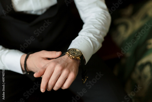 A man is wearing a black suit and a gold watch. He is sitting on a chair and looking at his watch