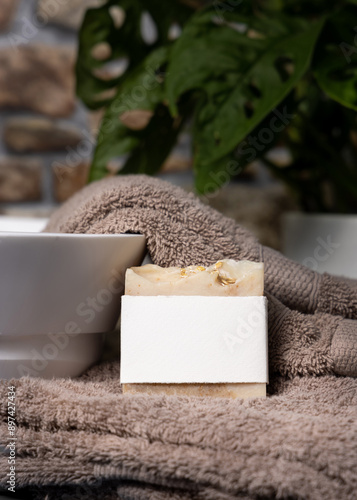 Soap bar with blank label on brown folded towels near plant and basin in bathroom, mockup photo