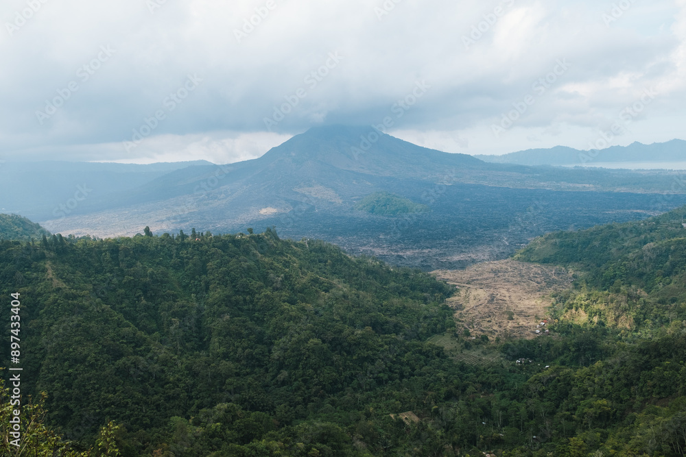 An expansive view of Mount Batur volcano with a lush green valley in the foreground, under a cloudy sky. An expansive view of Mount Batur volcano with a lush green valley in the foreground, under a cloudy sky.