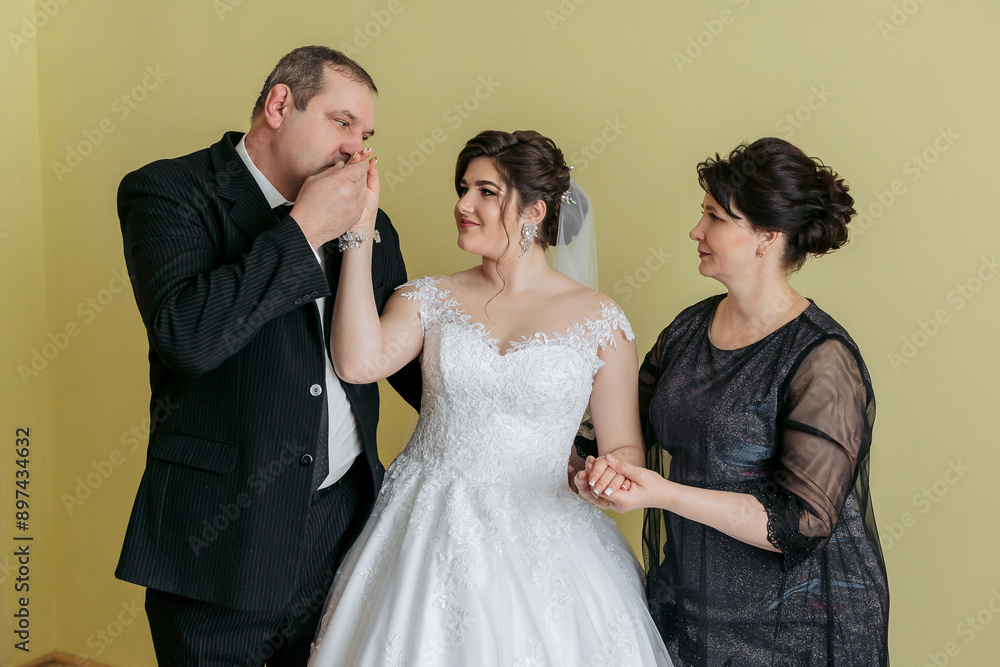 A man and a woman are hugging a young bride. The man is touching the bride's nose. The bride is wearing a white dress. The scene is a wedding