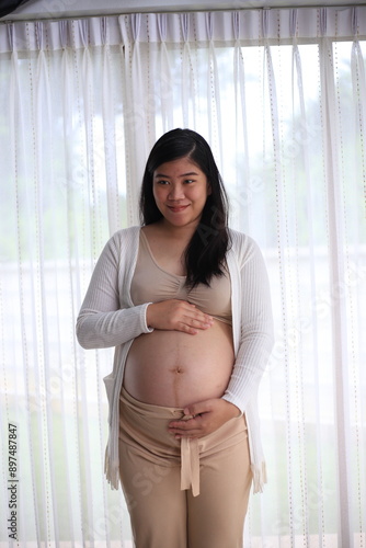 Happy pregnant woman with big tummy relax at home. Portrait of pregnant mature woman standing near the window and caresses her belly. Smiling woman expecting child and looking at camera. Free Download Happy pregnant woman with big tummy relax at home. Portrait of pregnant mature woman standing near the window and caresses her belly. Smiling woman expecting child and looking at camera. - GFXLoad
