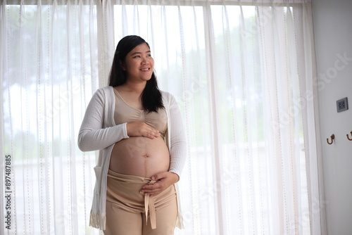 Happy pregnant woman with big tummy relax at home. Portrait of pregnant mature woman standing near the window and caresses her belly. Smiling woman expecting child and looking at camera. Free Download Happy pregnant woman with big tummy relax at home. Portrait of pregnant mature woman standing near the window and caresses her belly. Smiling woman expecting child and looking at camera. - GFXLoad
