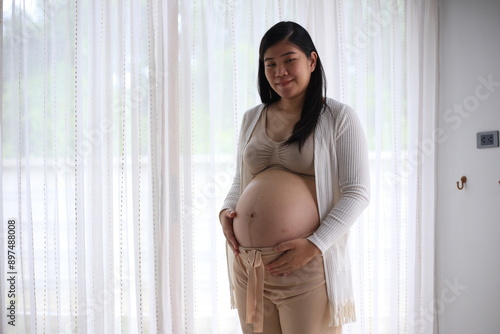 Happy pregnant woman with big tummy relax at home. Portrait of pregnant mature woman standing near the window and caresses her belly. Smiling woman expecting child and looking at camera. Free Download Happy pregnant woman with big tummy relax at home. Portrait of pregnant mature woman standing near the window and caresses her belly. Smiling woman expecting child and looking at camera. - GFXLoad