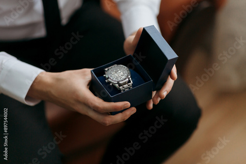 A man is holding a watch in a box. The watch is silver and black. The man is wearing a suit and tie. The watch is a gift for someone special