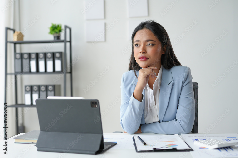 Young asian businesswoman is looking away and thinking about a new project while sitting at her desk in a modern office Young asian businesswoman is looking away and thinking about a new project while sitting at her desk in a modern office