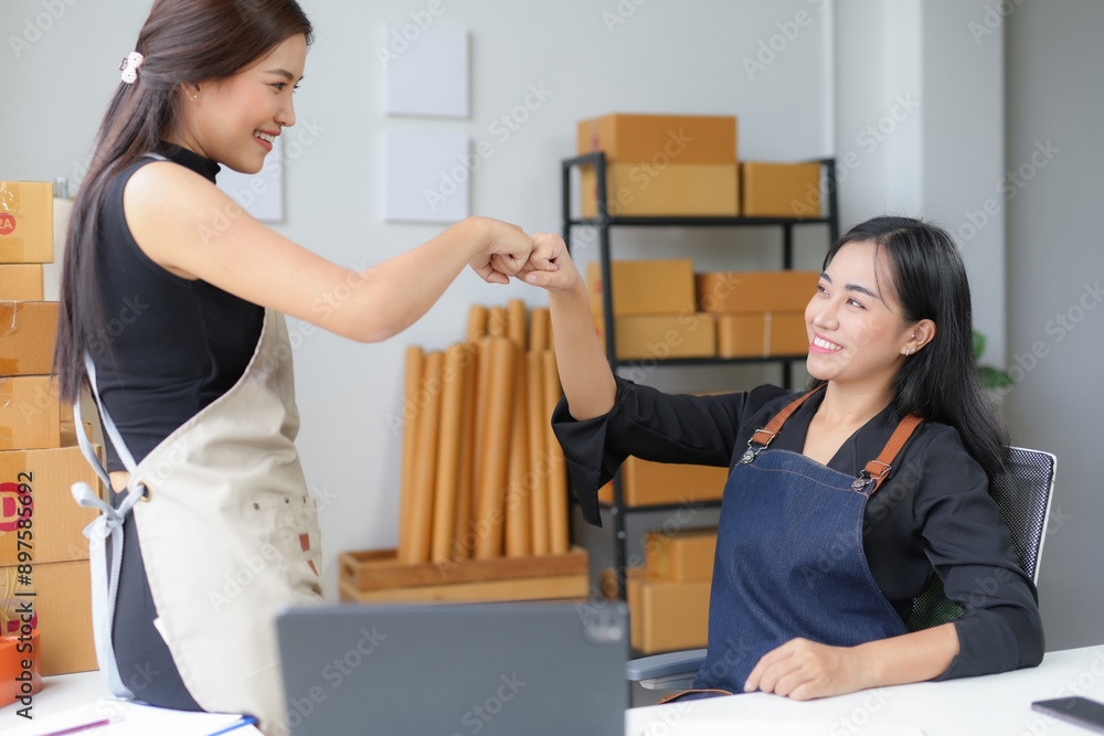 © amnaj - Two young women in a busy warehouse celebrate success with a fist bump, showing confidence and teamwork in their online business. Smiling and proud, they embody entrepreneurship and partnership