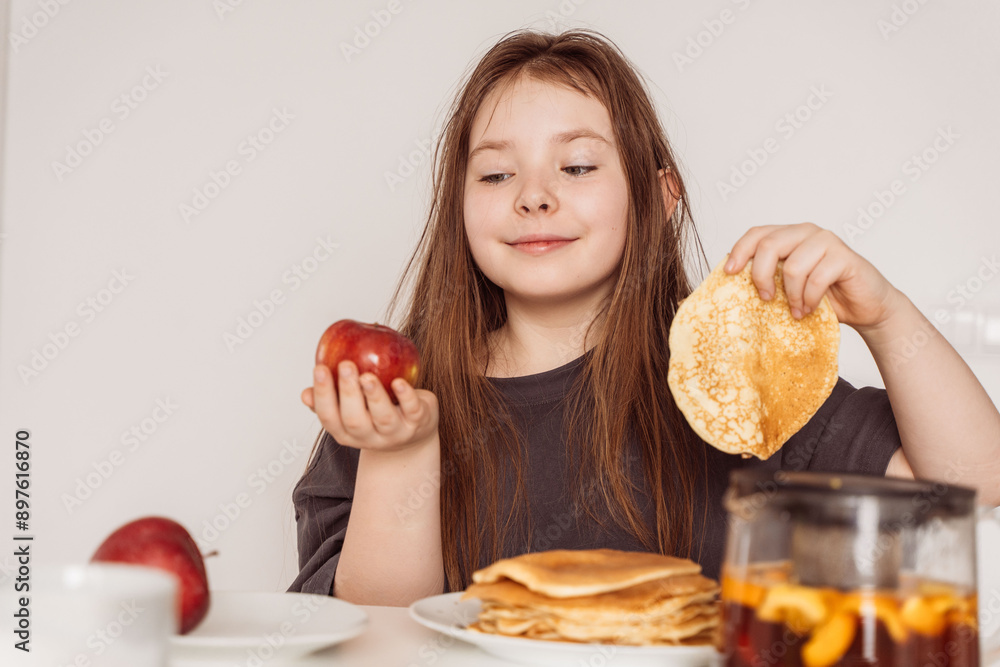 A little teenage girl holds a pancake in one hand and a red apple in the other and looks at it. Concept of choosing the right food A little teenage girl holds a pancake in one hand and a red apple in the other and looks at it. Concept of choosing the right food