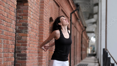 Fotografie A woman in a black tank top stretches with her arms behind her back, leaning against a brick wall