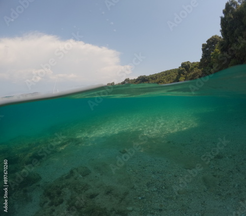 Over under, split level shot showing above and below the sea water. A bleached coral reef in shallow water with blue sky and green coastline above the water.