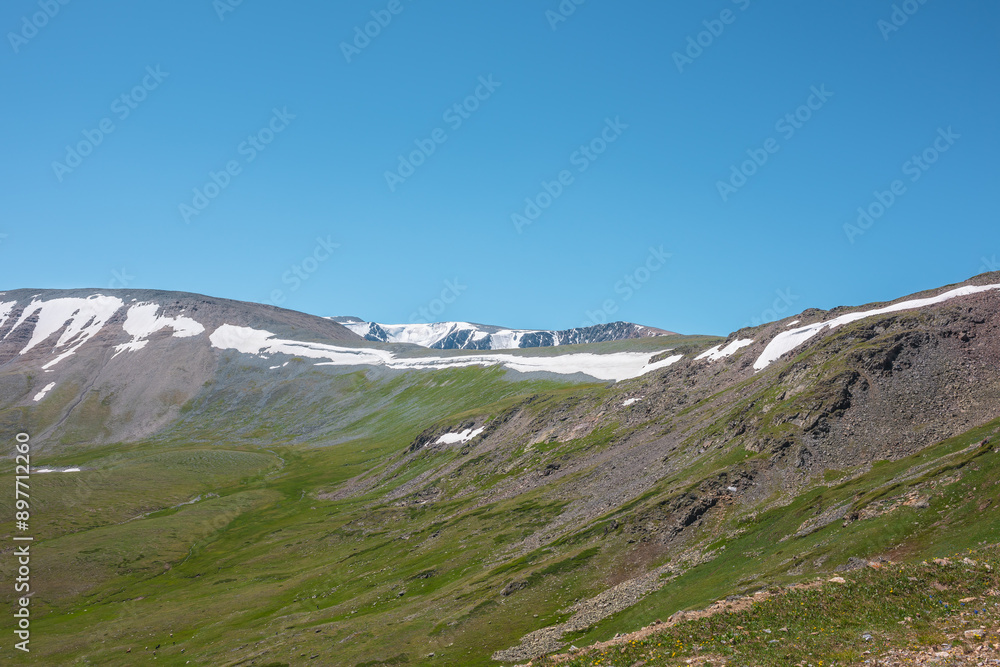 Fototapeta premium Vivid layered landscape with sunlit steep green rocky mountain wall with snow cornice against high snowy mountain range under clear blue sky. Glacier on top in bright sun. Sunny day in high mountains.